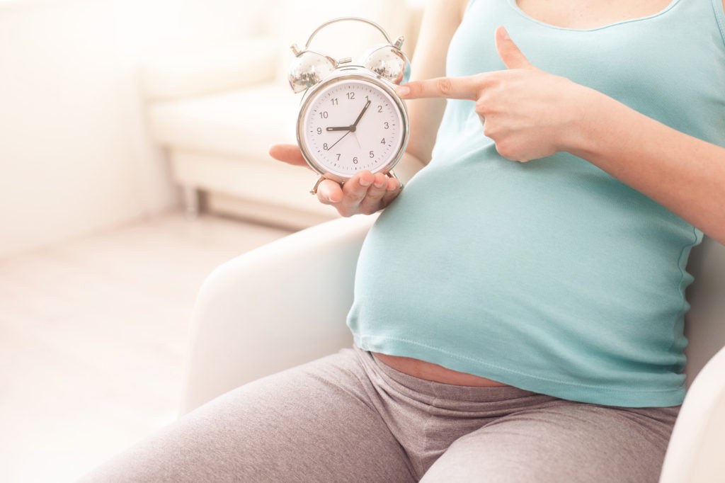 Cute pregnant woman is waiting for her child. She is sitting and pointing finger at clock