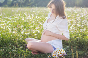 Happy pregnant woman is sitting in meadow of chamomile flowers, sunset time, new life concept