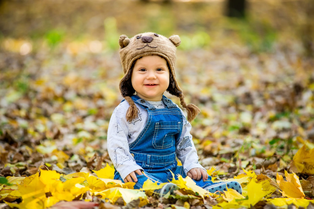 Happy, happy baby sitting in the park with yellow leaves in autumn in bear cap. In autumn park kid sitting in yellow leaves. Cheerful baby sitting in the park in bear cap. Warm autumn day in the park.